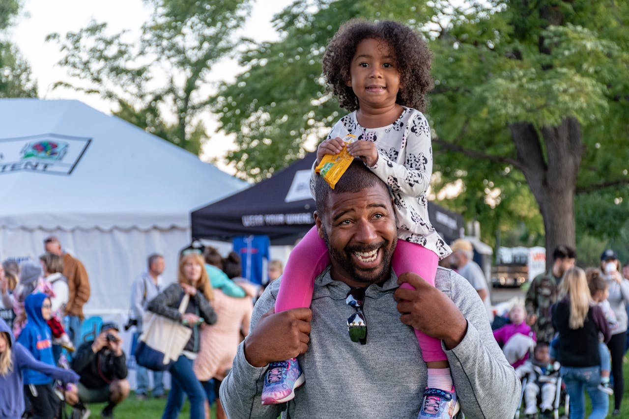 Young girl seated on father's shoulders at outdoor fair>